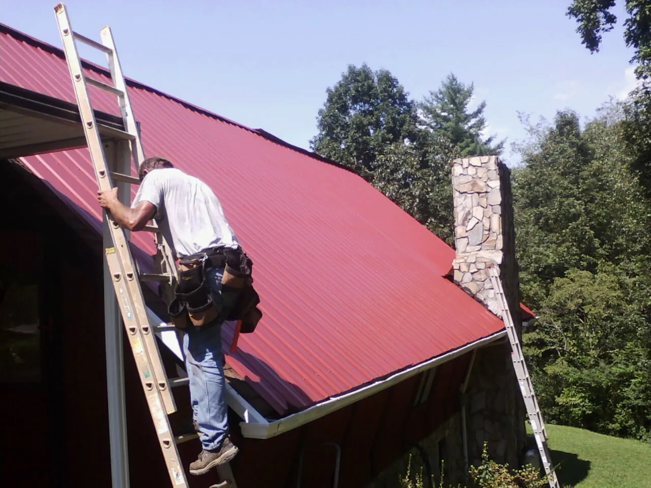 Roofer on a ladder beside a red metal roof and stone chimney — Soffit & Fascia Repair by Roofing Brothers in Piney Creek